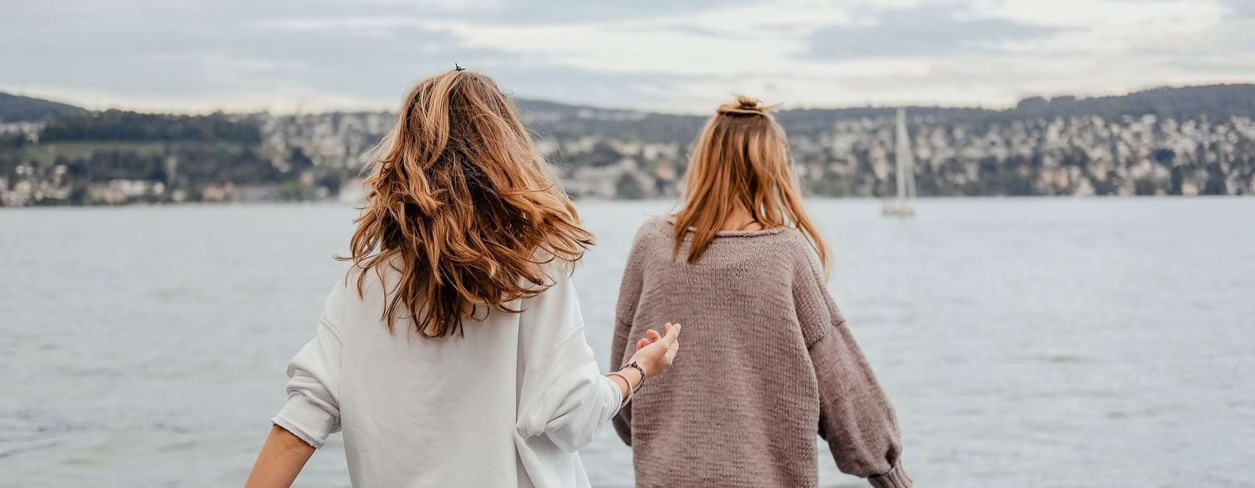 two women walk to the edge of a dock, in a body of water, looking toward the city on the other shore